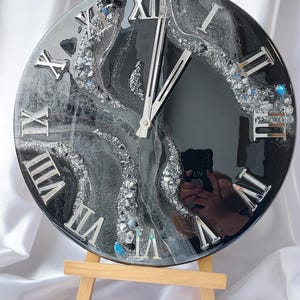 May include: A round, black and silver resin clock with silver Roman numerals. The clock face has a swirling design with embedded silver and blue accents. The clock is displayed on a small wooden easel.