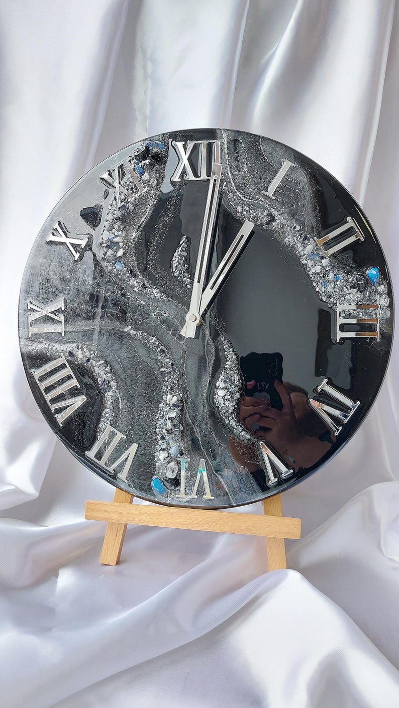 May include: A round, black and silver resin clock with silver Roman numerals. The clock face has a swirling design with embedded silver and blue accents. The clock is displayed on a small wooden easel.