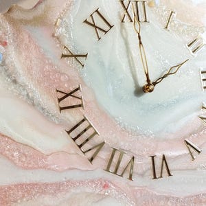 May include: A decorative wall clock with a unique, abstract design. The clock face features gold Roman numerals and hands against a backdrop of swirling white, pink, and gold resin. The clock is mounted on a wooden easel.
