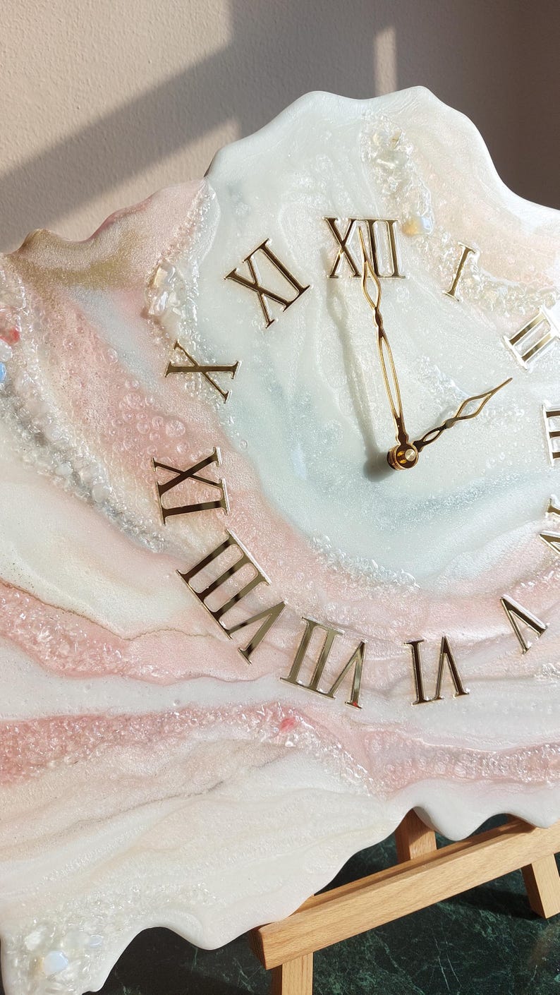 May include: A decorative wall clock with a unique, abstract design. The clock face features gold Roman numerals and hands against a backdrop of swirling white, pink, and gold resin. The clock is mounted on a wooden easel.