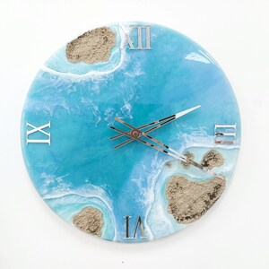 May include: A blue and white resin clock with Roman numeral markings and a beach scene design. The clock face features white waves and brown sand islands.