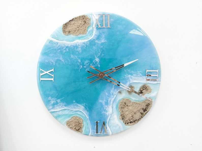 May include: A blue and white resin clock with Roman numeral markings and a beach scene design. The clock face features white waves and brown sand islands.