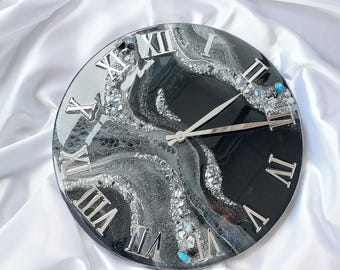 May include: Round wall clock with a black and silver resin design. The clock features silver Roman numerals and hands. The face has a marbled effect with embedded crystals and turquoise accents. A decorative piece for home decor.