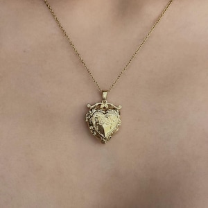 May include: A gold-toned heart-shaped locket pendant suspended from a delicate matching gold-toned chain. The locket features an intricate embossed floral and scrollwork design on its front surface, with a small decorative bail at the top.