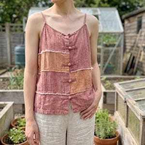 Puede incluir: Una camiseta de tirantes con diseño de patchwork en tonos rosa empolvado y naranja apagado. La prenda tiene tirantes finos, escote en pico y una tapeta frontal con cuatro cierres de palanca cubiertos de tela y costuras deshilachadas.