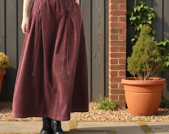 May include: A burgundy corduroy skirt with a wide, flowing silhouette. The skirt features drawstring details on the front and a comfortable elastic waistband. The model is wearing black boots. The skirt is knee-length.