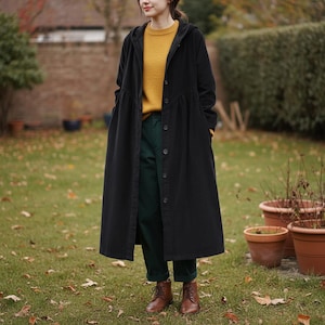 Könnte beinhalten: Ein langer schwarzer Mantel mit Kapuze und Knopfleiste. Der Mantel wird über einem gelben Pullover und einer grünen Hose getragen. Braune Stiefel vervollständigen das Outfit. Die Person steht in einer grasbewachsenen Fläche mit Herbstlaub und Blumentöpfen.