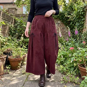 Könnte beinhalten: Weit geschnittene Cordhose in tiefem Burgunderrot mit elastischem Bund, zwei Seitentaschen und dekorativen schwarzen Kordelzügen auf der Vorderseite. Die Hose besteht aus einem strukturierten Stoff mit vertikalen Rippen und hat einen lockeren Schnitt.