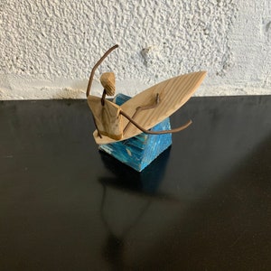 Op de afbeelding: Een houten sculptuur van een surfer op een golf. De surfer is gemaakt van licht hout en draad, met een surfboard en een blauwe blokbasis. De sculptuur staat tegen een witte, getextureerde muur en een zwart oppervlak.