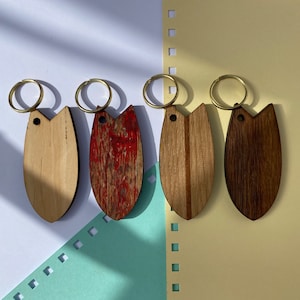 Könnte beinhalten: Vier Holz-Schlüsselanhänger in Surfbrettform mit Messingringen. Die Schlüsselanhänger sind in verschiedenen Farben erhältlich, darunter Naturholz, verwittertes Rot und Dunkelbraun. Sie sind auf einem farbenfrohen Hintergrund angeordnet.