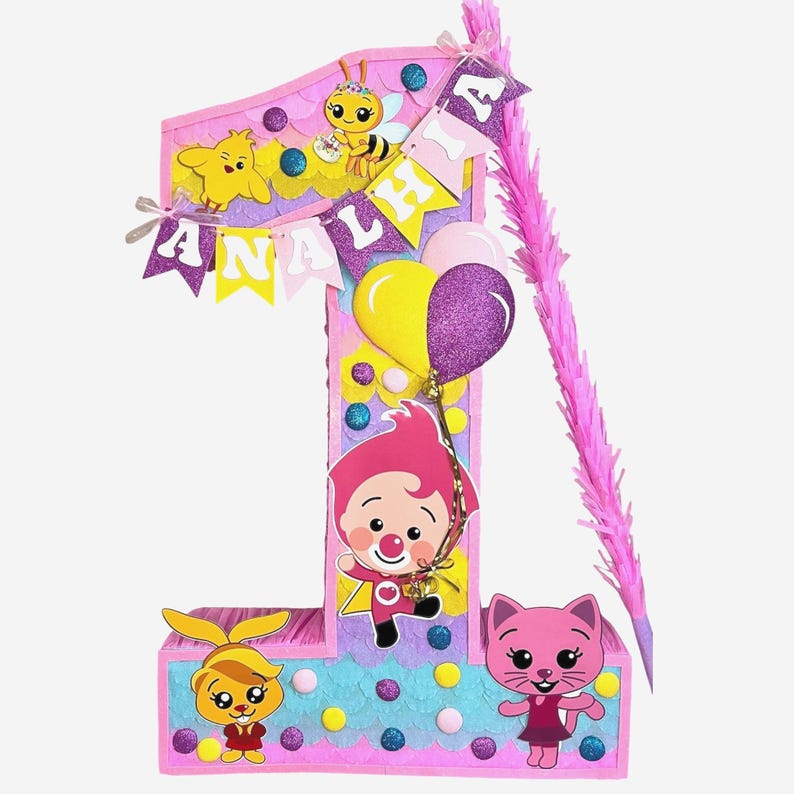 May include: A number one shaped pinata in shades of pink, blue, and yellow. It features cartoon character cutouts, a banner with the name ANALHIA, and two balloons. A pink fringe stick is attached to the side, decorated with glittery accents.