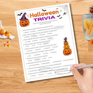 May include: A Halloween trivia game sheet with a pumpkin graphic and the title "Halloween Trivia." The sheet includes multiple-choice questions about Halloween. A hand is circling the correct answers with a pen. Candy corn and a decorative drink are also present.