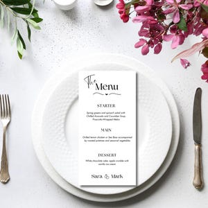 May include: A wedding menu card with the text "The Menu" in elegant script, placed on a white plate with a fork and knife. The menu lists starter, main, and dessert options. Floral arrangements and greenery surround the plate.