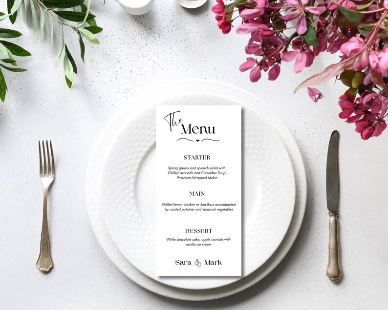 May include: A wedding menu card with the text "The Menu" in elegant script, placed on a white plate with a fork and knife. The menu lists starter, main, and dessert options. Floral arrangements and greenery surround the plate.