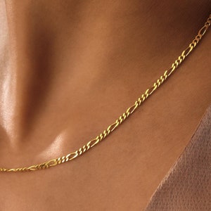 Op de afbeelding: Een delicate goudkleurige Figaro-ketting. De ketting is om de nek gedrapeerd. De ketting heeft een herhalend patroon van drie kleine schakels, gevolgd door één langere schakel. De achtergrond is effen bruin.