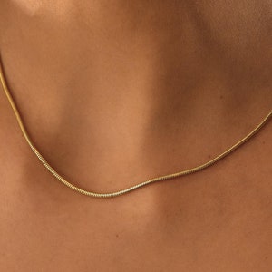May include: A close-up of a woman wearing a delicate gold chain necklace. The chain is thin and has a subtle shine. The woman's skin is smooth and her hands are gently touching the necklace.