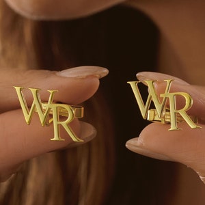 Könnte beinhalten: Ein Paar goldene Manschettenknöpfe mit den eingravierten Buchstaben "WR". Die Manschettenknöpfe werden von den Fingern einer Person gehalten.