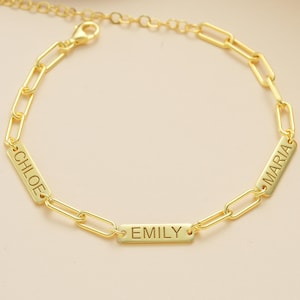 Puede incluir: Una pulsera de cadena dorada con placas rectangulares con nombres. Los nombres "CHLOE", "EMILY" y "MARIA" están grabados. La pulsera está sobre una superficie dorada.