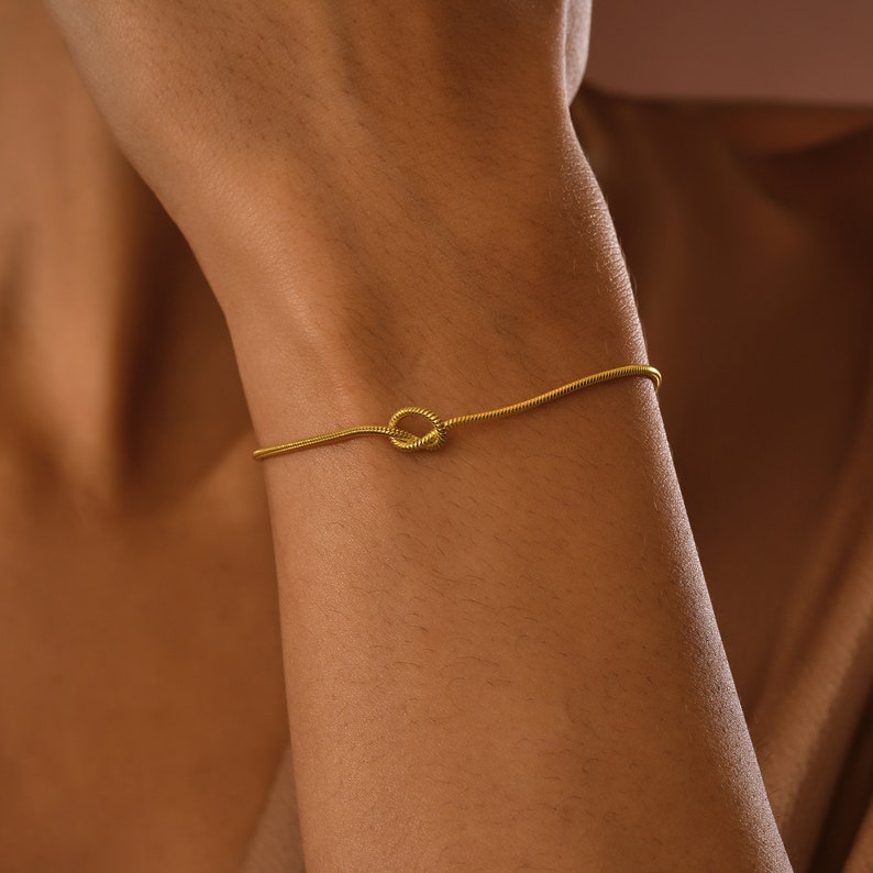 Peut inclure: Un bracelet en cha&icirc;ne dor&eacute;e avec un motif de n&oelig;ud. Le bracelet est port&eacute; au poignet.