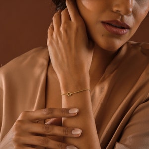 Peut inclure: Un bracelet d&eacute;licat en or avec un design simple de n&oelig;ud. Le bracelet est pr&eacute;sent&eacute; sur le poignet d'une personne.
