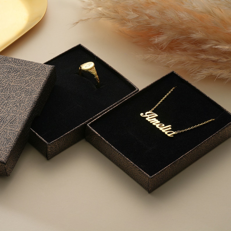May include: Two small, black, velvet-lined boxes with gold jewelry inside. One box contains a gold ring, the other contains a gold necklace with the name "Amelia" on it.