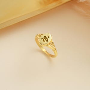 Könnte beinhalten: Ein goldener Siegelring mit ovaler Oberfläche, auf der der Buchstabe "S" in dunkler Farbe abgebildet ist. Der Ring hat ein verziertes Filigranmuster am Band und um die Oberfläche. Der Ring ist vor einem weichen, neutralen Hintergrund platziert.