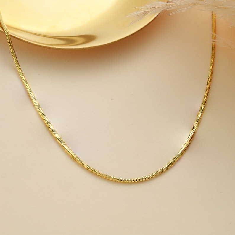May include: A gold snake chain necklace on a light beige background.