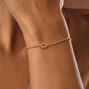 Peut inclure: Un bracelet en cha&icirc;ne dor&eacute;e avec un motif de n&oelig;ud. Le bracelet est port&eacute; au poignet.