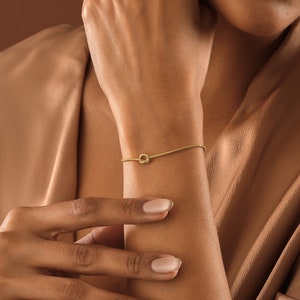 Peut inclure: Un bracelet en or avec un n&oelig;ud sur le poignet d'une personne. Le bracelet est fin et d&eacute;licat, avec un design simple de n&oelig;ud.