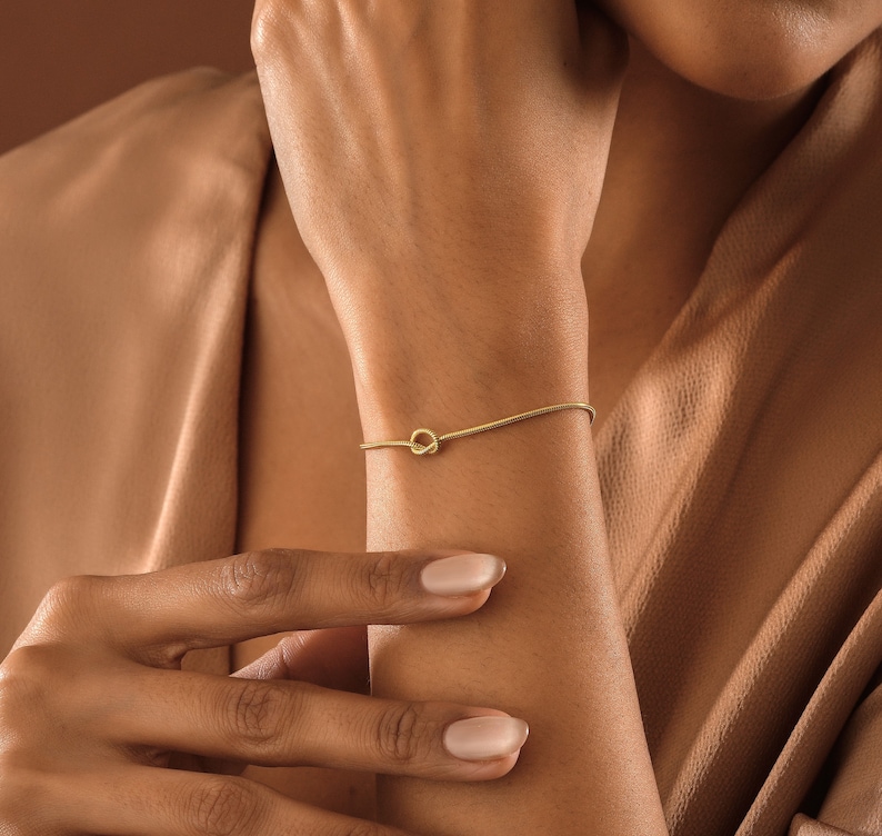 Peut inclure: Un bracelet en or avec un n&oelig;ud sur le poignet d'une personne. Le bracelet est fin et d&eacute;licat, avec un design simple de n&oelig;ud.