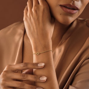 Peut inclure: Un bracelet en or avec un n&oelig;ud sur le poignet d'une personne. Le bracelet est fin et d&eacute;licat, avec un design simple de n&oelig;ud.