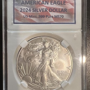 May include: A silver American Eagle coin in a plastic case. The coin features the image of Lady Liberty with the words "In God We Trust" and "2024". The label on the case reads "American Eagle 2024 Silver Dollar US Mint .999 Pure MS70".
