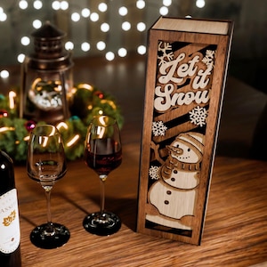 Könnte beinhalten: Eine festliche Urlaubsszene mit einer Weinflasche, zwei Weingläsern und einer Holzkiste mit einem Schneemann-Design und dem Schriftzug "Let it Snow". Eine brennende Kerze und ein dekorativer Kranz sind ebenfalls vorhanden.