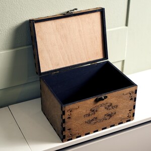May include: An open wooden box with a black interior and a decorative design on the front. The box is made of light brown wood and has a hinged lid.