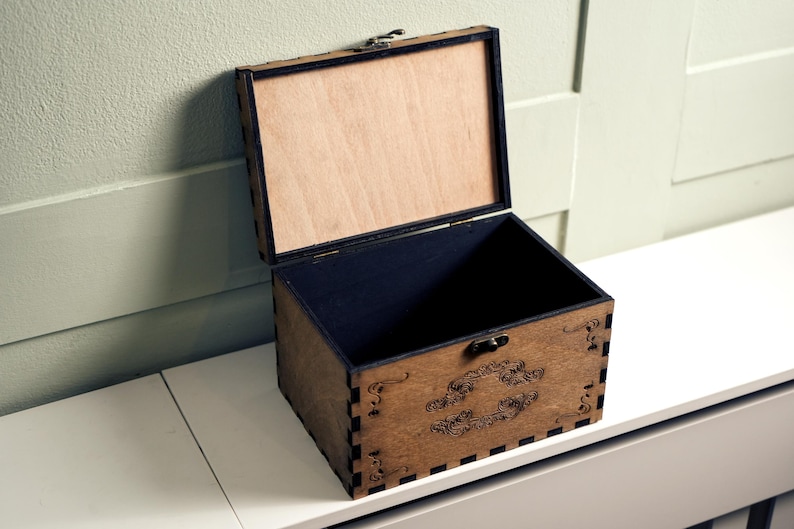 May include: An open wooden box with a black interior and a decorative design on the front. The box is made of light brown wood and has a hinged lid.