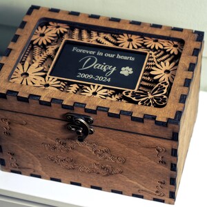 May include: A wooden box with a glass top and a laser-cut design of flowers and butterflies. The box has a brass latch and a plaque that reads "Forever in our hearts Daisy 2009-2024".