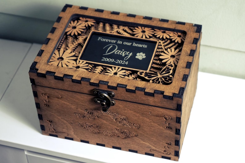 May include: A wooden box with a glass top and a laser-cut design of flowers and butterflies. The box has a brass latch and a plaque that reads "Forever in our hearts Daisy 2009-2024".