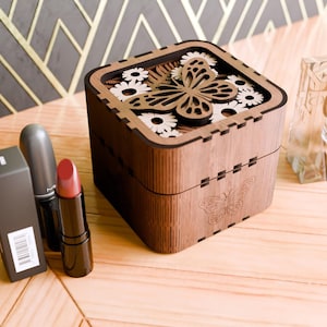 May include: A wooden box with a butterfly design and floral accents. The box is made of dark wood and has a light brown finish. The butterfly is cut out of the wood and has a natural wood colour. The flowers are also cut out of the wood and have a white finish.