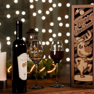 May include: A Christmas-themed wine gift set featuring a dark wine bottle, two wine glasses, and a wooden box with a reindeer design and "Merry Christmas" text. A lit candle and string lights are in the background.