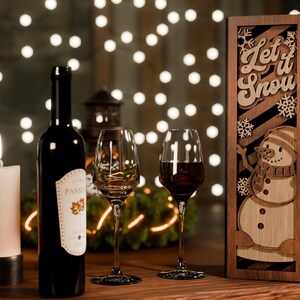May include: A festive scene with a wine bottle, two wine glasses, and a wooden wine box. The box features a snowman design and the words "Let it Snow." A lit candle and string lights add to the holiday ambiance.