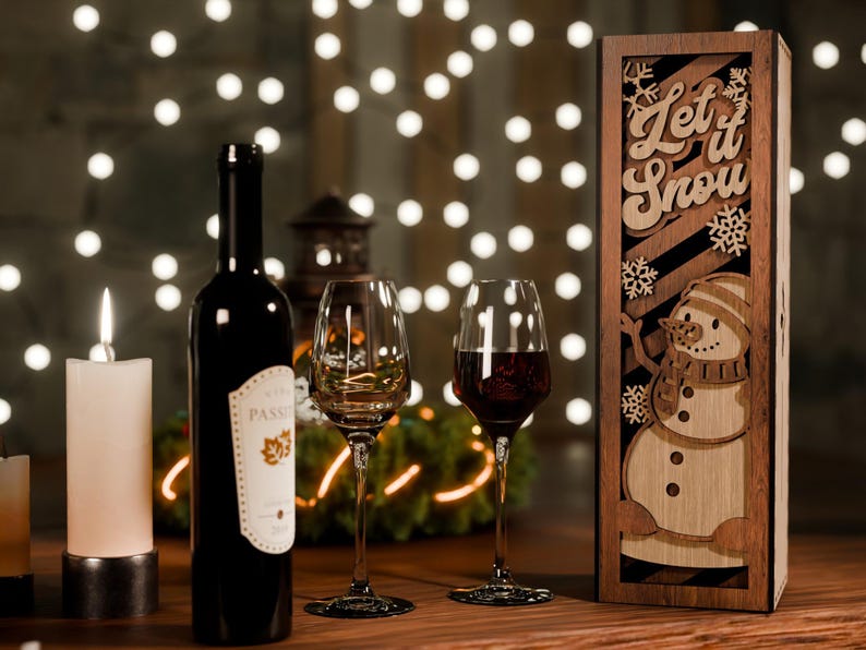 May include: A festive scene with a wine bottle, two wine glasses, and a wooden wine box. The box features a snowman design and the words "Let it Snow." A lit candle and string lights add to the holiday ambiance.