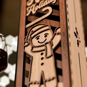 May include: Wooden wine bottle holder with a gingerbread man design. The holder has a light brown finish with a dark brown accent. The words "Happy Holidays" are carved at the top. A wine glass with red wine is in the foreground.