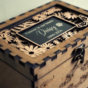May include: A wooden box with a hinged lid and a latch. The lid features a laser-cut design of flowers and ferns. A small plaque on the lid reads "Forever in our hearts Daisy 2009-2024".