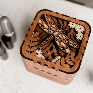 May include: A wooden box featuring a laser-cut design of a dragonfly and flowers. The box is crafted from dark wood and has a natural finish.