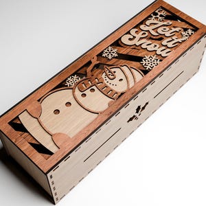 May include: Wooden rectangular box with a laser-cut design featuring a snowman, snowflakes, and the words "Let it Snow." The box is made of light and dark brown wood, with a decorative winter theme.