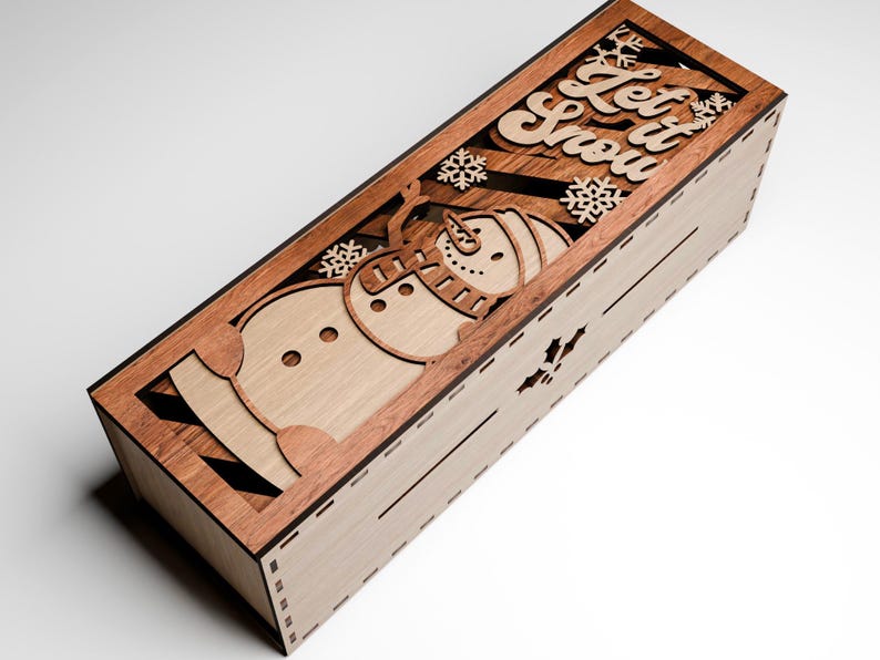 May include: Wooden rectangular box with a laser-cut design featuring a snowman, snowflakes, and the words "Let it Snow." The box is made of light and dark brown wood, with a decorative winter theme.