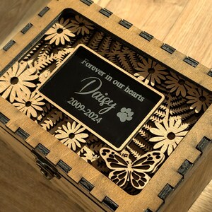 May include: Wooden box with a laser-cut design of flowers and a butterfly. The box has a black plaque with the text "Forever in our hearts Daisy 2009-2024" and a paw print.