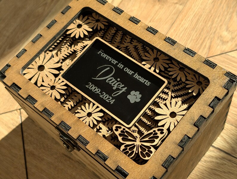 May include: Wooden box with a laser-cut design of flowers and a butterfly. The box has a black plaque with the text "Forever in our hearts Daisy 2009-2024" and a paw print.