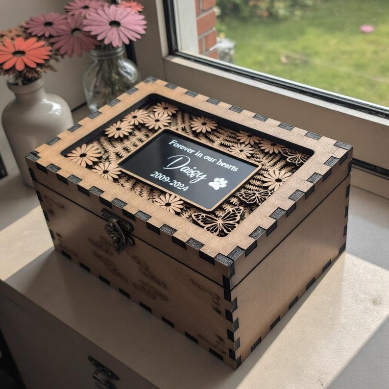 May include: A wooden box with a cutout design of flowers and butterflies. The box has a black plaque with the text "Forever in our hearts Daisy 2009-2024" and a paw print.
