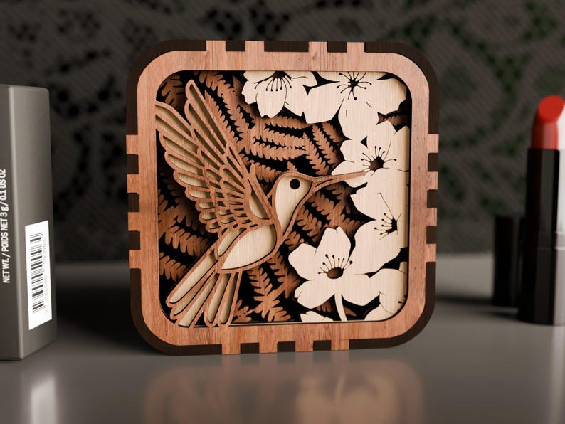 May include: A wooden box featuring a laser-cut design of a hummingbird in flight, surrounded by flowers and ferns. The box is brown with a dark brown frame.